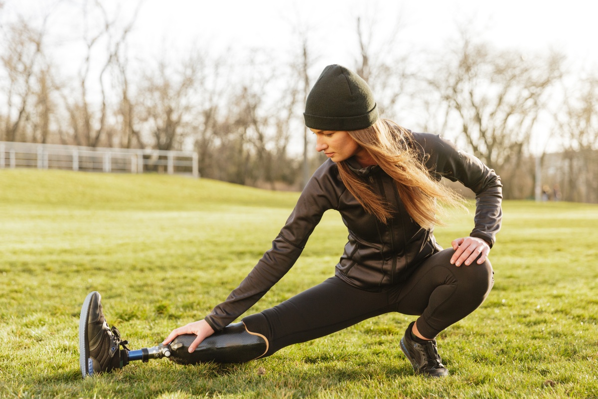 A women is stretching on grass wearing fall layers on and has a prosthetic leg.